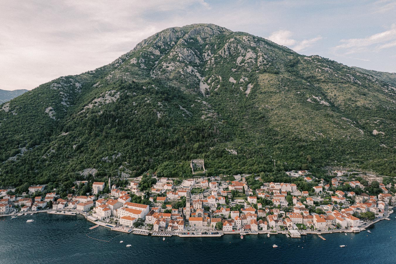 Aerial view of a coastal town with red-roofed buildings nestled at the base of a lush, green mountain, bordered by a calm blue sea.