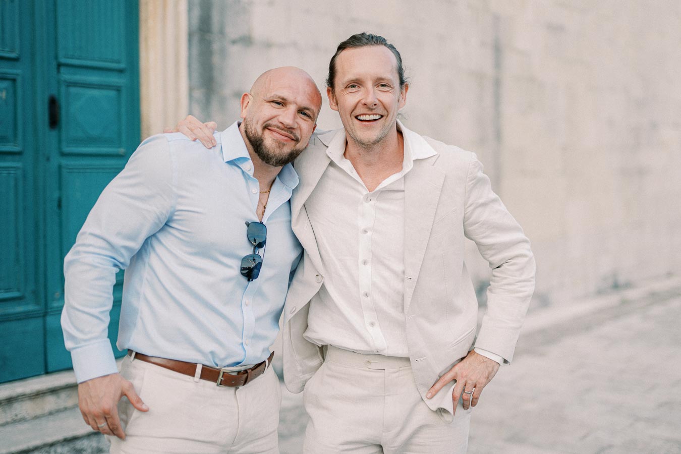 Two men smiling and posing together in light-colored outfits, one in a light blue shirt with sunglasses hanging from the collar, standing outdoors in front of a blue door and stone wall.