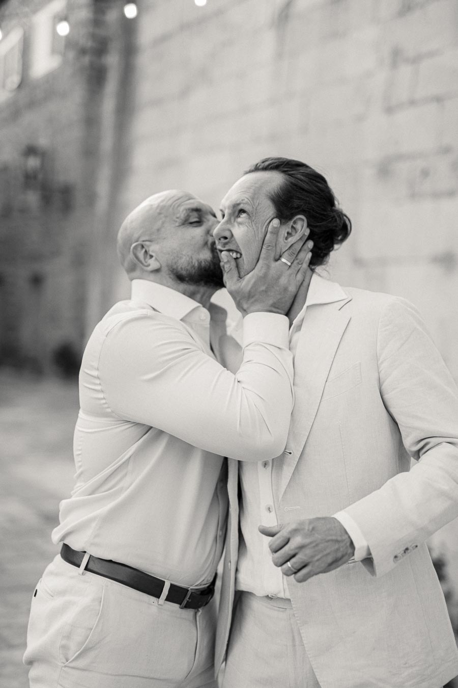 Two men in suits sharing a joyful, affectionate moment in a black-and-white photo, with one playfully kissing the other on the cheek, conveying friendship and happiness.