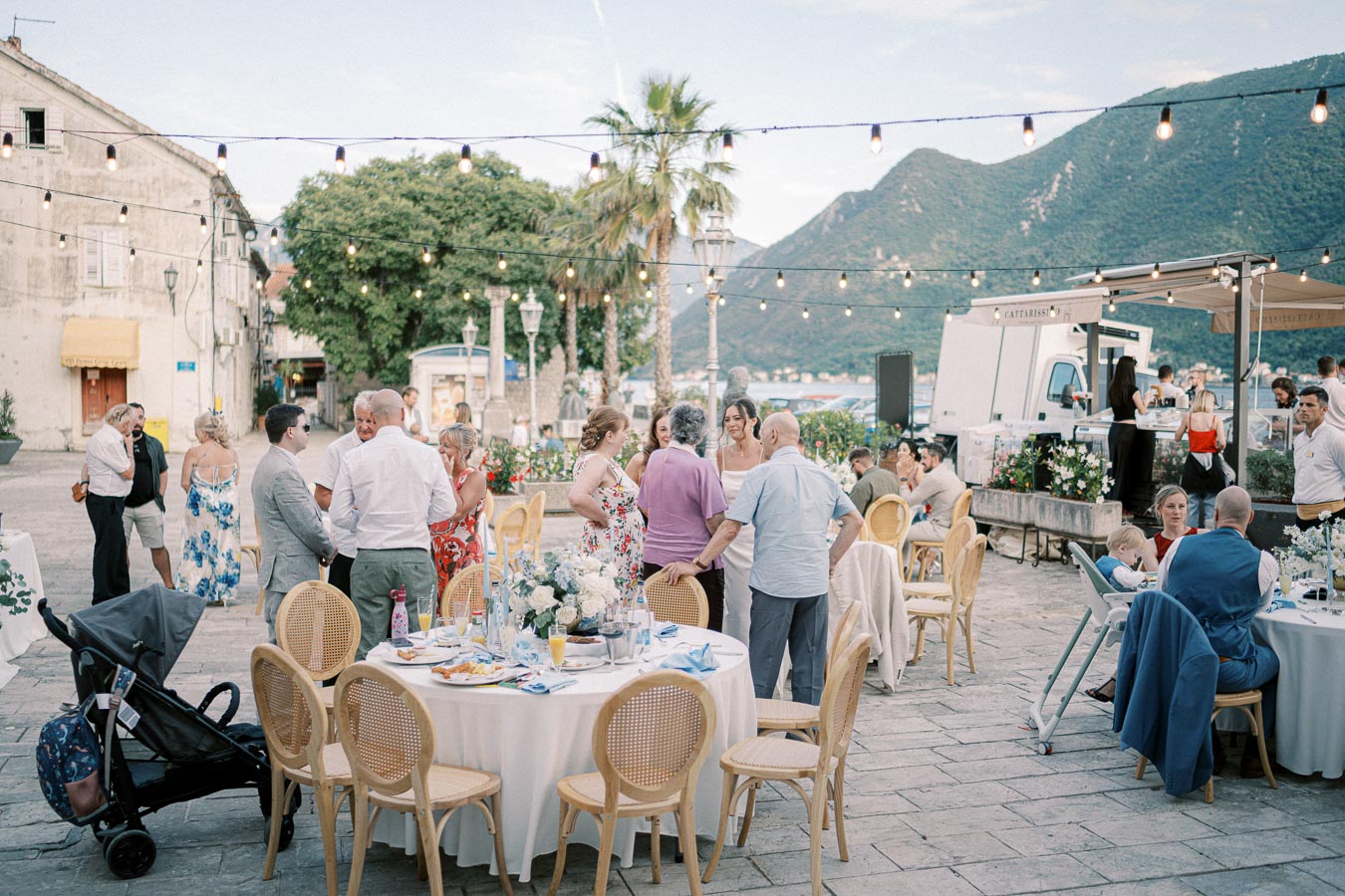 Outdoor wedding reception with guests socializing around tables on a picturesque patio, featuring string lights and mountain views.