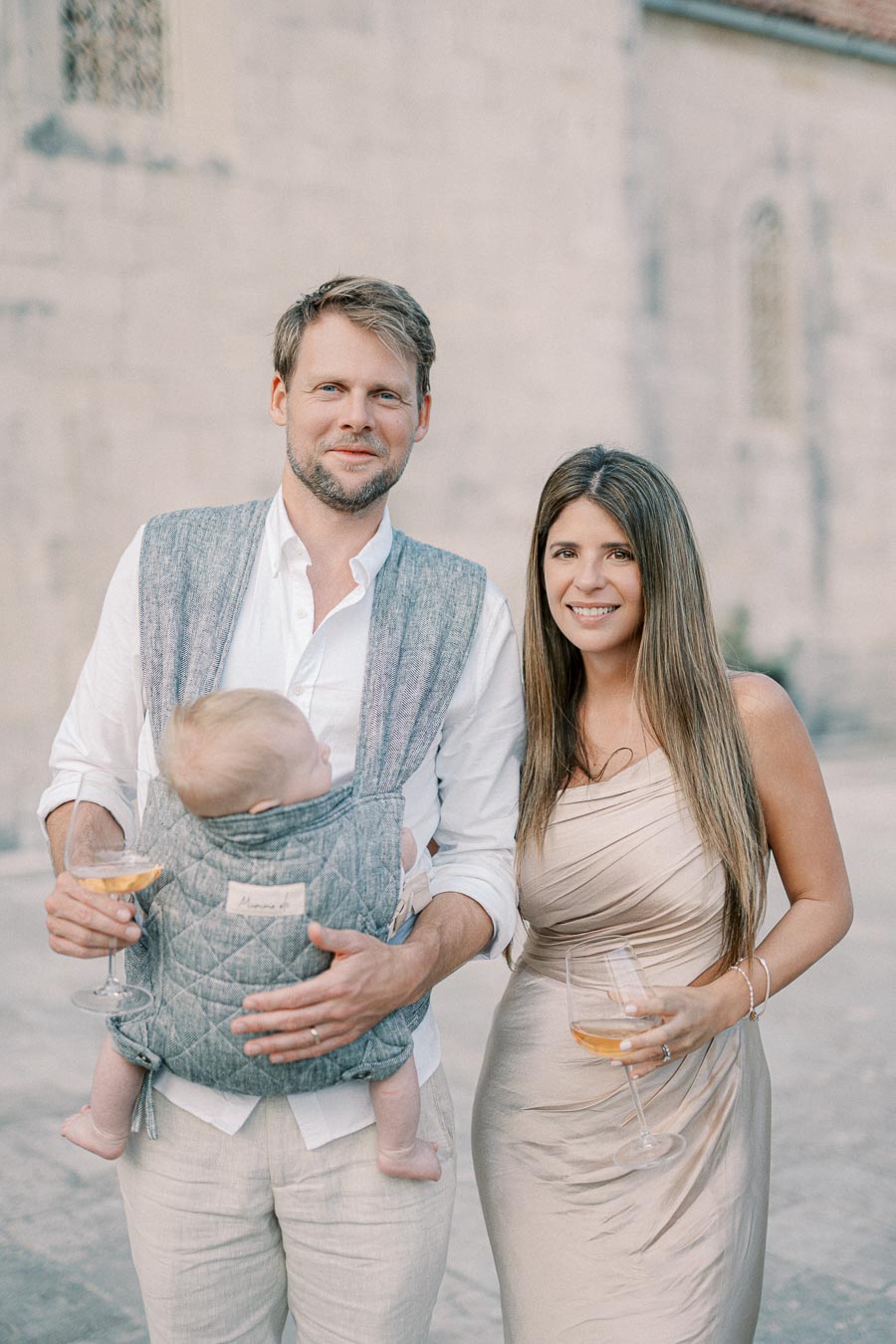 A couple enjoying a celebration with a baby carrier holding an infant. The man is holding a wine glass, dressed in casual chic attire, standing next to a woman in an elegant beige dress, both smiling outdoors.
