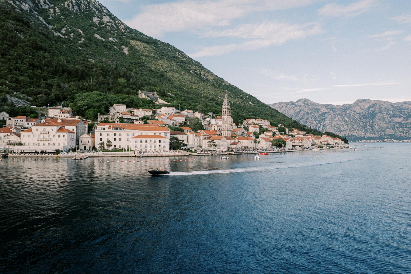 Scenic coastal town with traditional red-roofed buildings nestled against green hills and mountains, overlooking calm blue waters and a speeding boat.