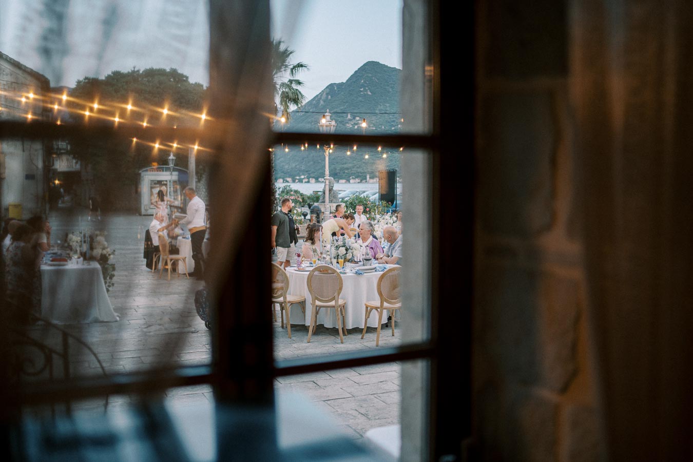 Outdoor evening wedding reception with guests seated at elegantly decorated tables under string lights, framed by a window with a view of mountains and a serene waterfront in the background.