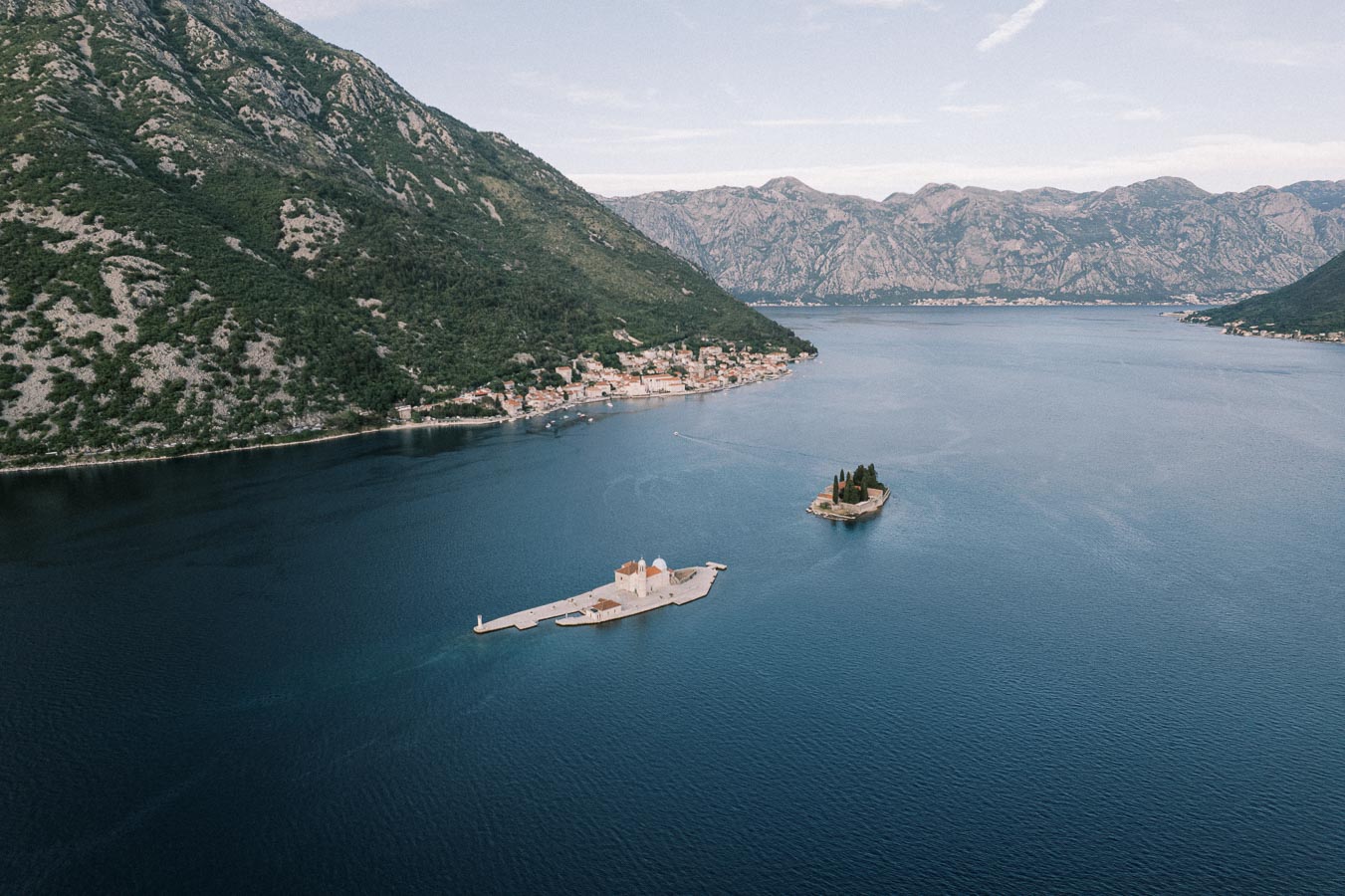Aerial view of the scenic Bay of Kotor in Montenegro, featuring two small islands with historic buildings surrounded by tranquil blue waters and mountainous terrain.