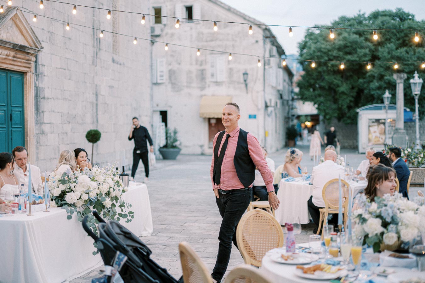 Outdoor wedding reception with elegantly dressed guests seated at decorated tables under string lights, set against a backdrop of historic stone buildings.