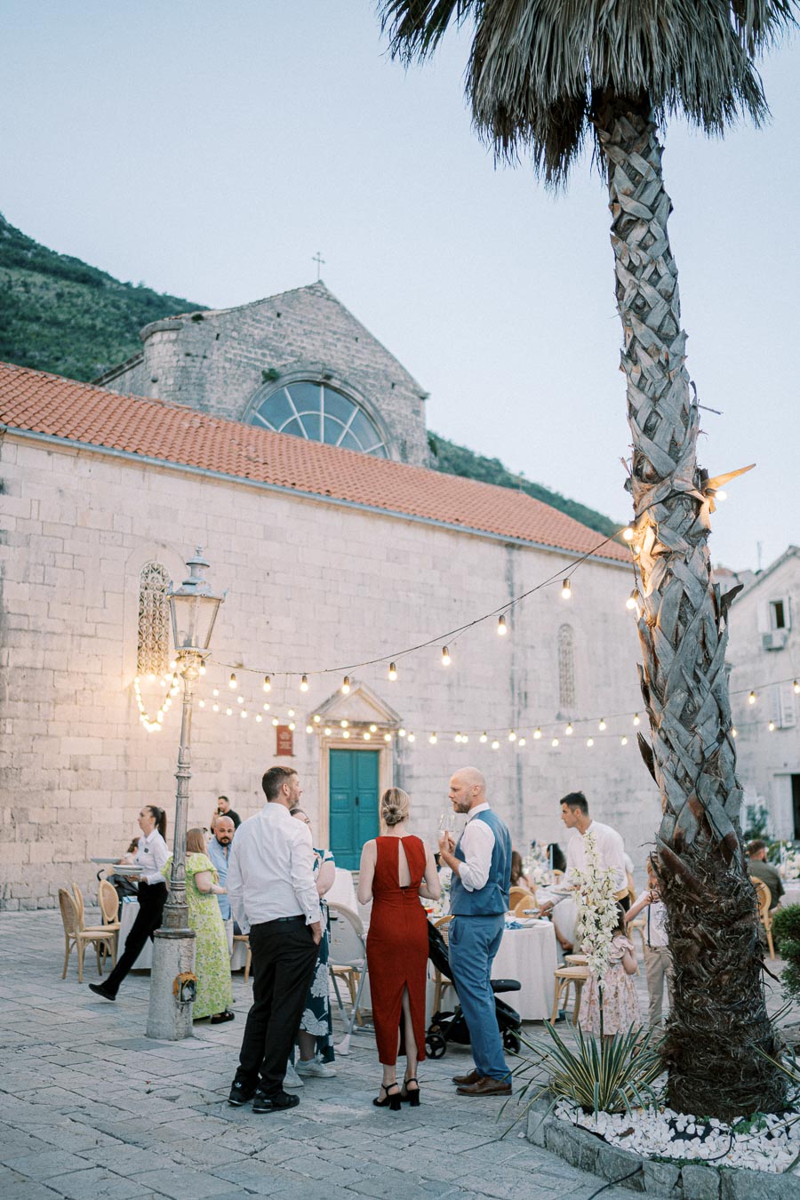 Elegant outdoor wedding reception in a historic stone courtyard with string lights and palm tree, featuring guests in formal attire enjoying the evening atmosphere.