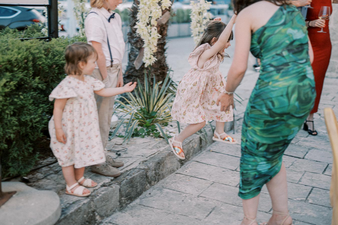 Children playing outdoors at a family gathering, with one child jumping off a small ledge while adults watch nearby.