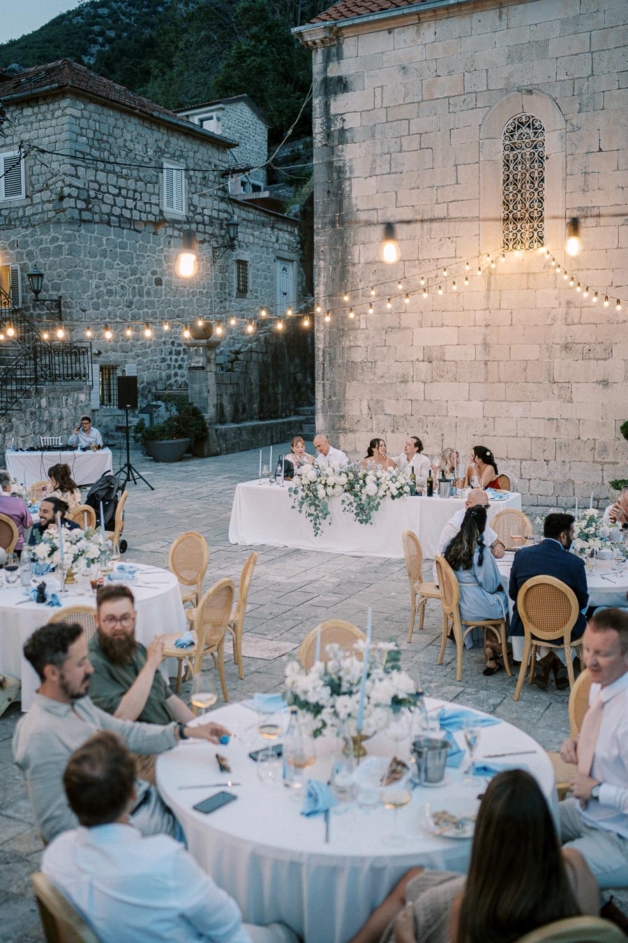 Evening outdoor wedding reception with guests seated at round tables adorned with floral arrangements, under string lights in a charming stone courtyard setting.