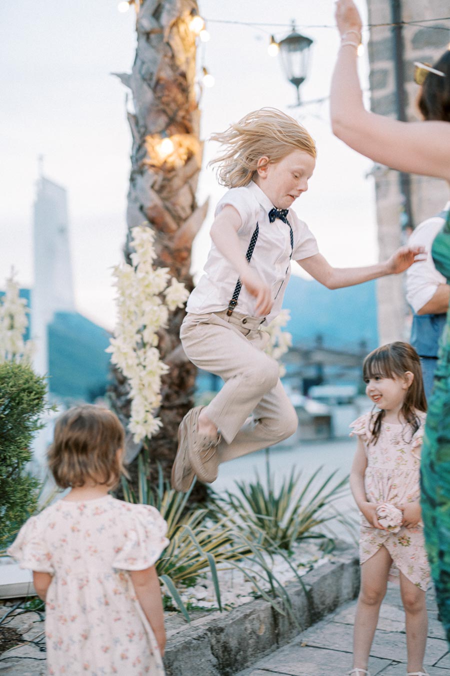 A joyful child in a white shirt and bow tie leaps into the air during an outdoor party, surrounded by two young girls in floral dresses, near palm trees and decorative lights.