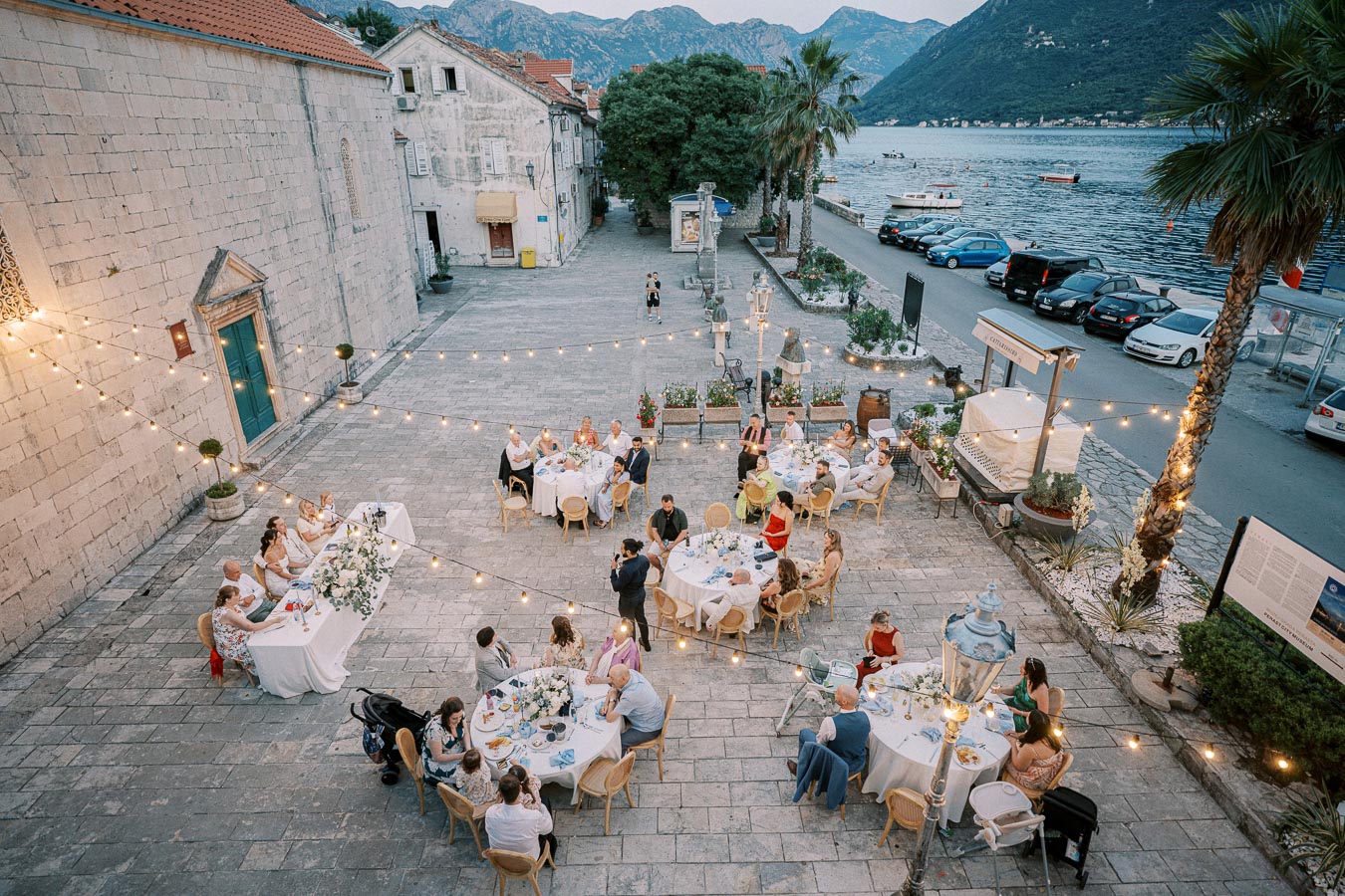 Aerial view of an outdoor wedding reception near a waterfront, featuring string lights, guests seated at round tables with white tablecloths, and a scenic mountain backdrop.