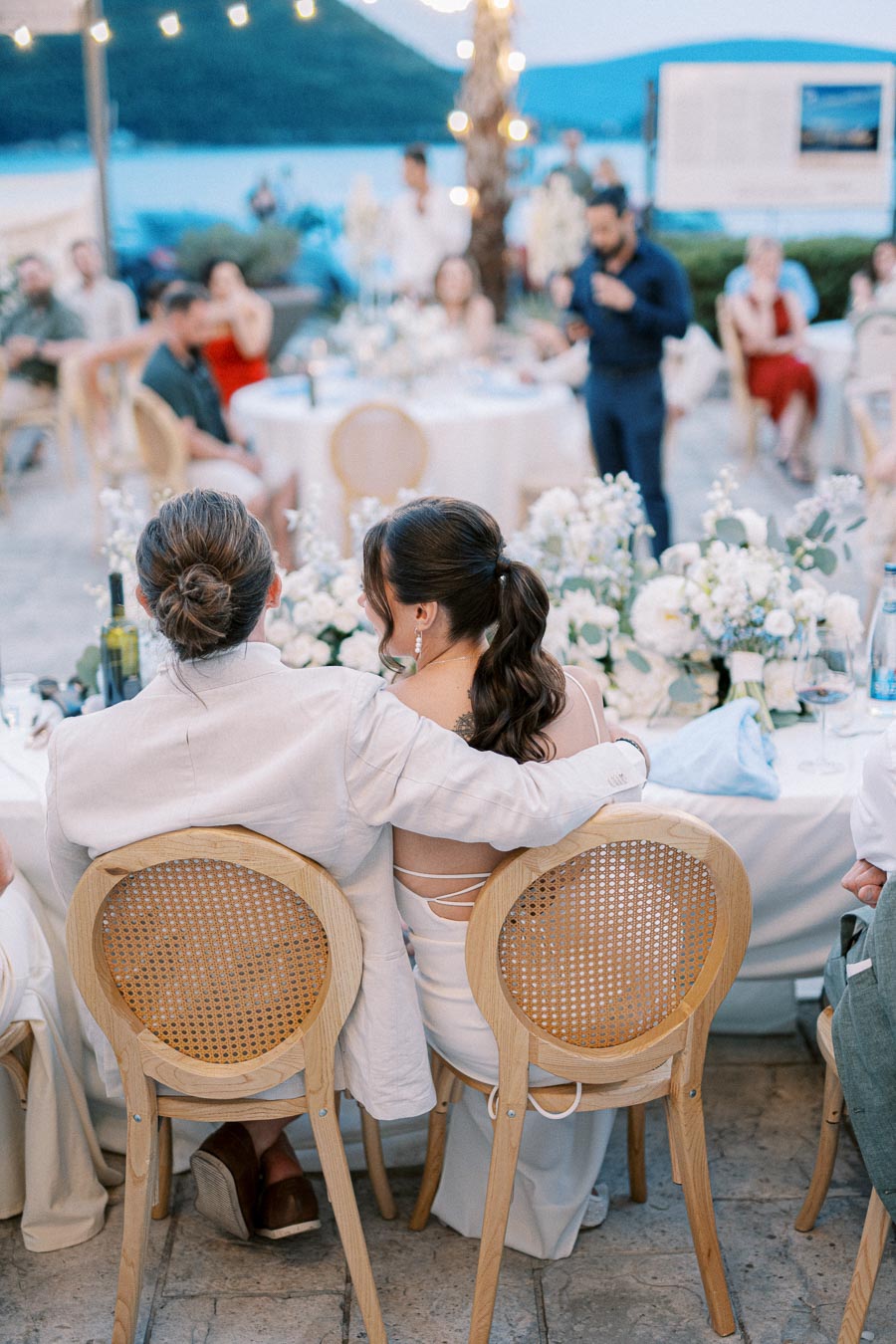A couple sitting closely together at an outdoor wedding reception, surrounded by beautifully arranged floral decorations and guests, with a scenic view of the mountains and a lake in the background.