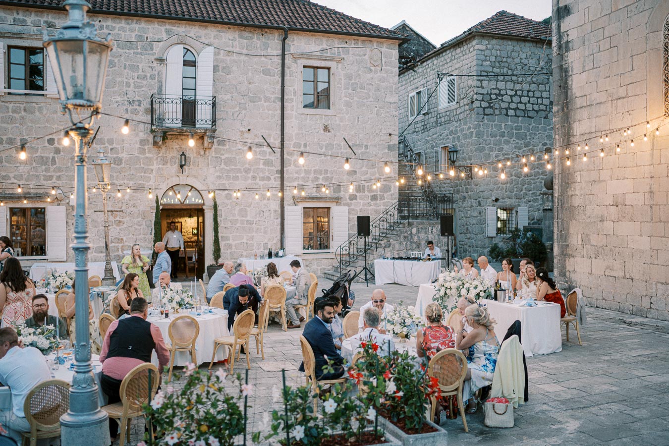 Outdoor evening wedding reception in a rustic courtyard with stone buildings and string lights, guests seated at round tables adorned with floral arrangements.