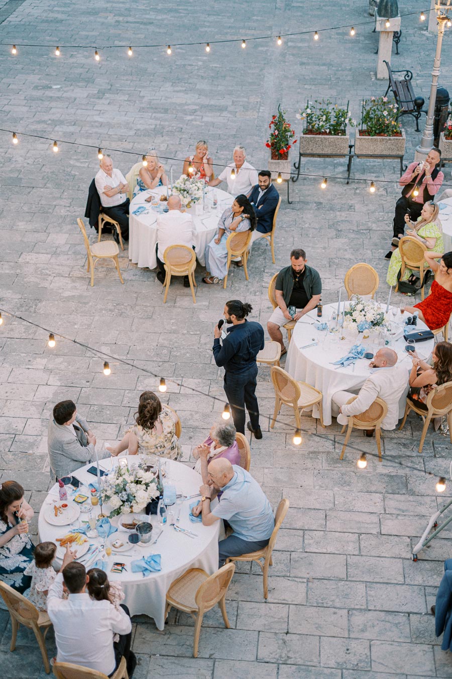 Guests seated around tables at an outdoor event with string lights, enjoying a formal gathering, while a speaker addresses the audience.