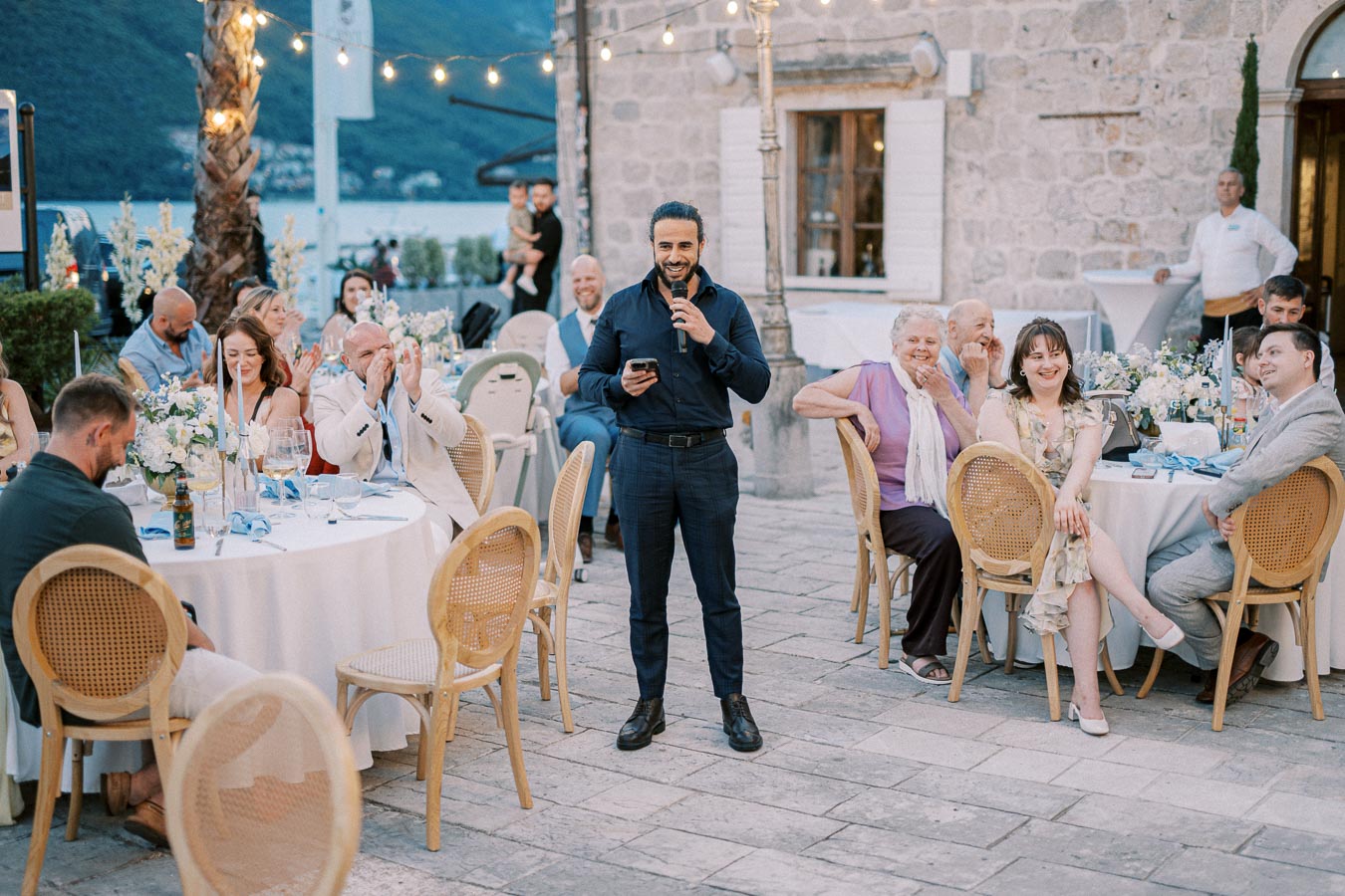 A man giving a speech with a microphone at an outdoor wedding reception, surrounded by seated guests at round tables. The setting features string lights, floral centerpieces, and a scenic view of distant hills by the water.