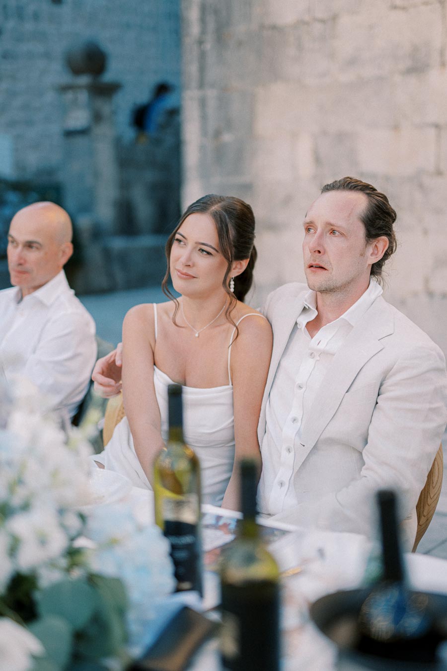 A couple in formal attire sits at an elegantly decorated table during an outdoor event. The woman is wearing a white dress and the man is in a light-colored suit. There are wine bottles and floral arrangements on the table, set against a stone wall backdrop.