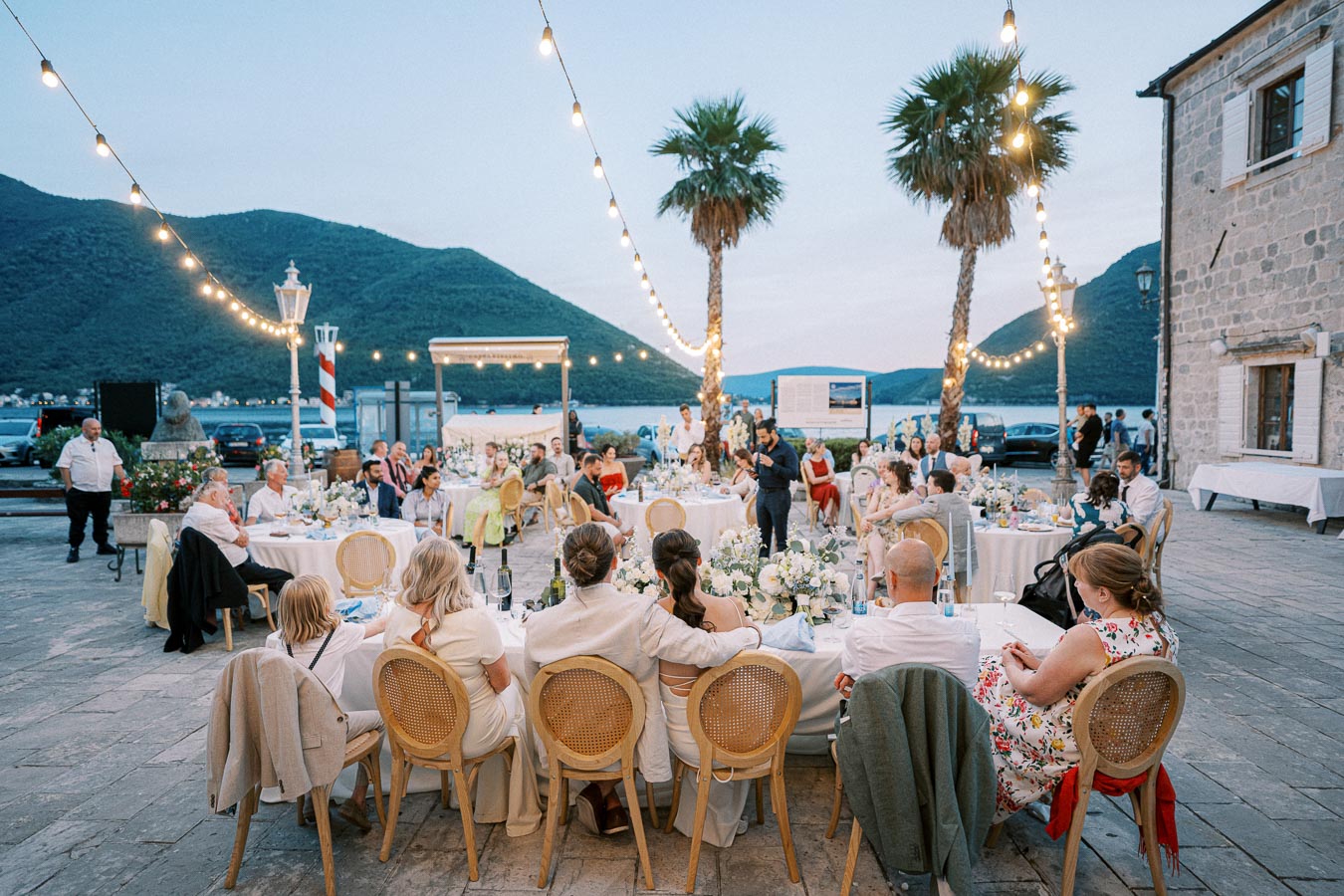 Outdoor wedding reception by the sea at sunset with guests seated around round tables, decorated with string lights and palm trees, against a backdrop of mountains and waterfront views.