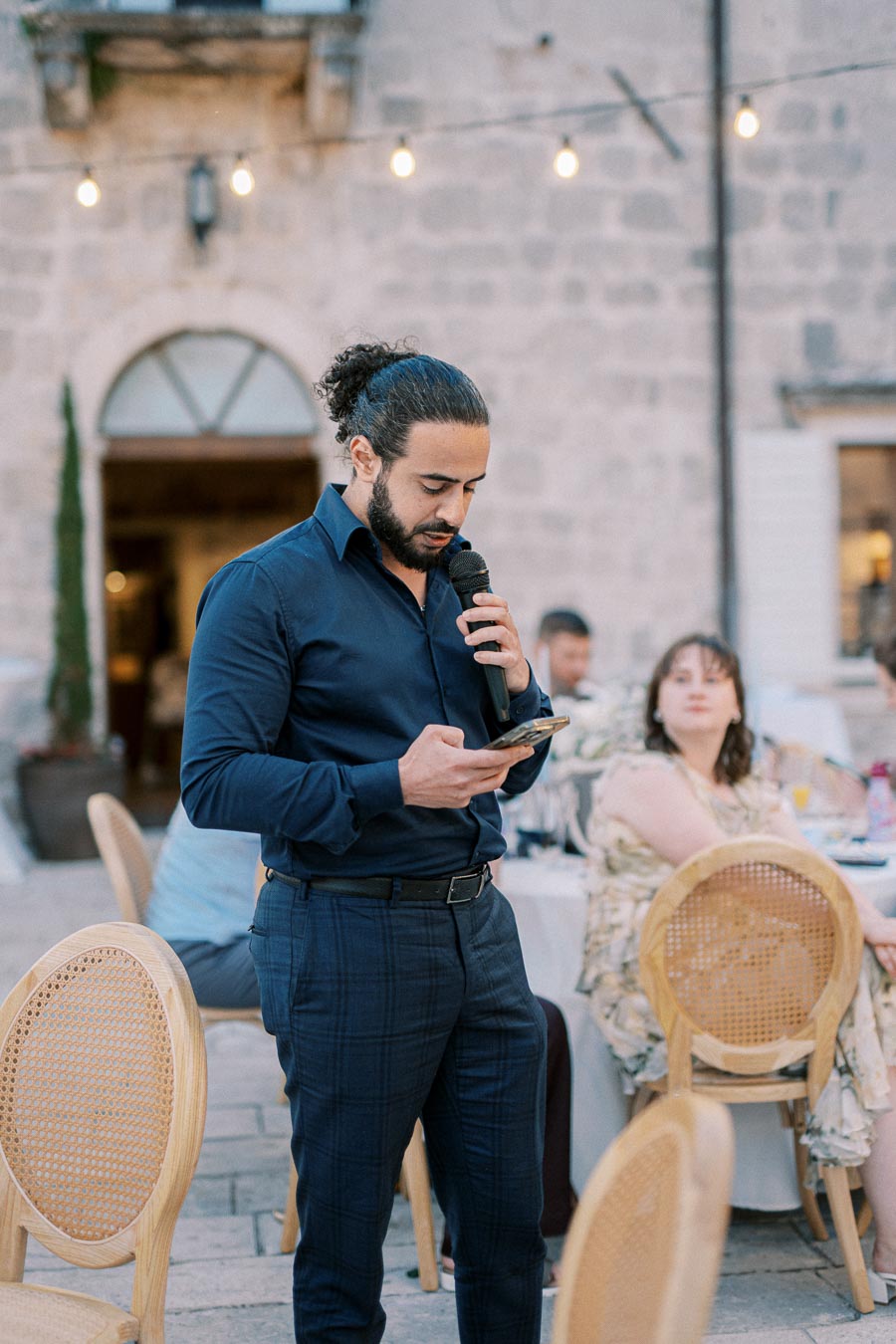 A man in a blue shirt speaks into a microphone while reading from a smartphone at an outdoor event, with guests seated at tables in the background and decorative lights hanging overhead.