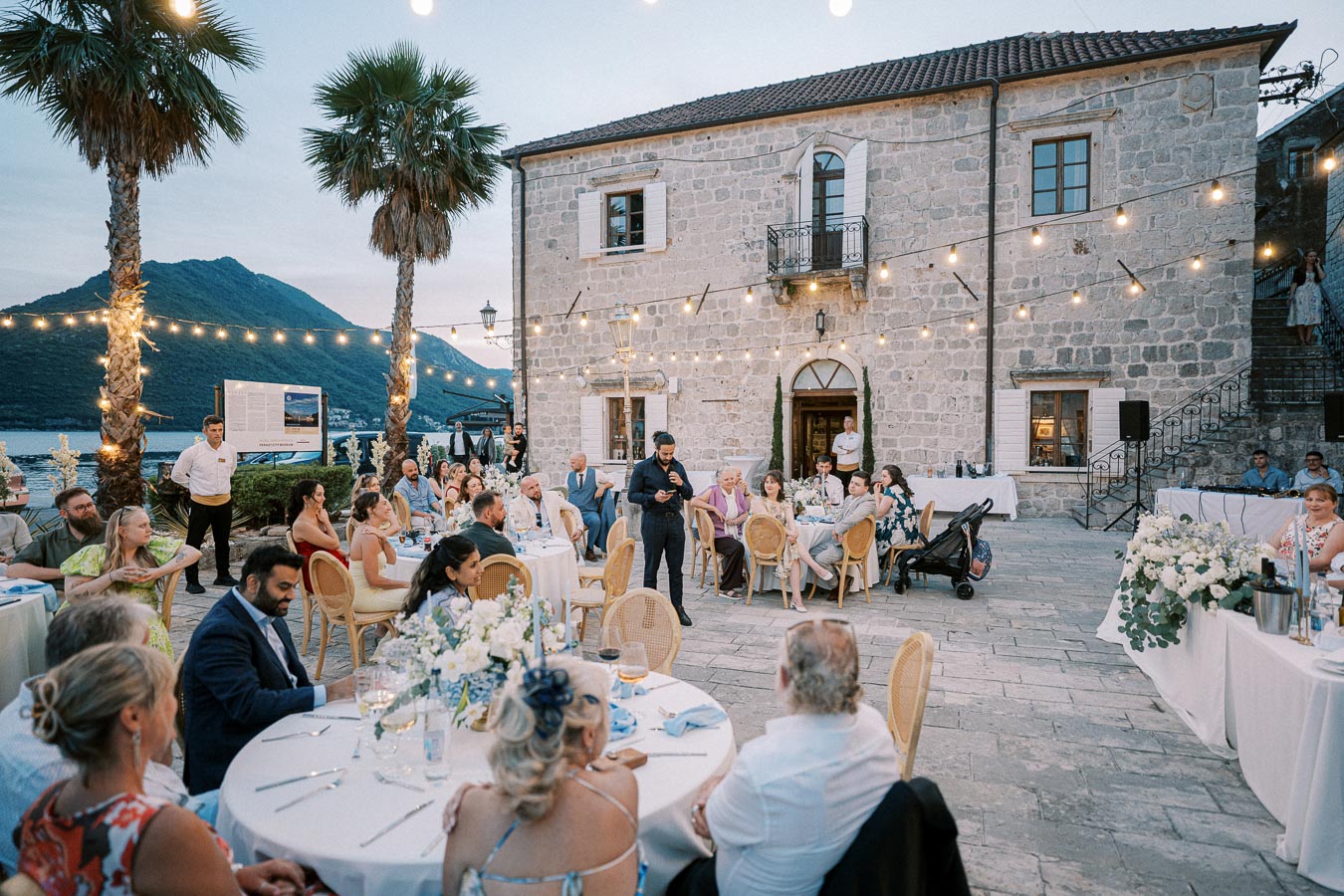 Outdoor wedding reception at a scenic venue, featuring guests seated at round tables adorned with floral centerpieces. The stone building and string lights create a romantic ambiance. Mountains and waterfront visible in the background enhance the picturesque setting.