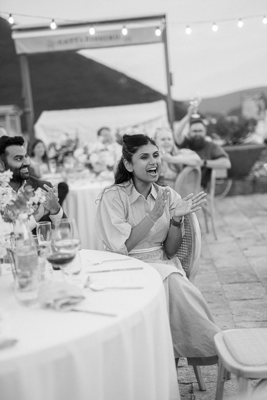 Black and white photo of people enjoying an outdoor event, with a woman clapping at a decorated table amid a festive atmosphere.