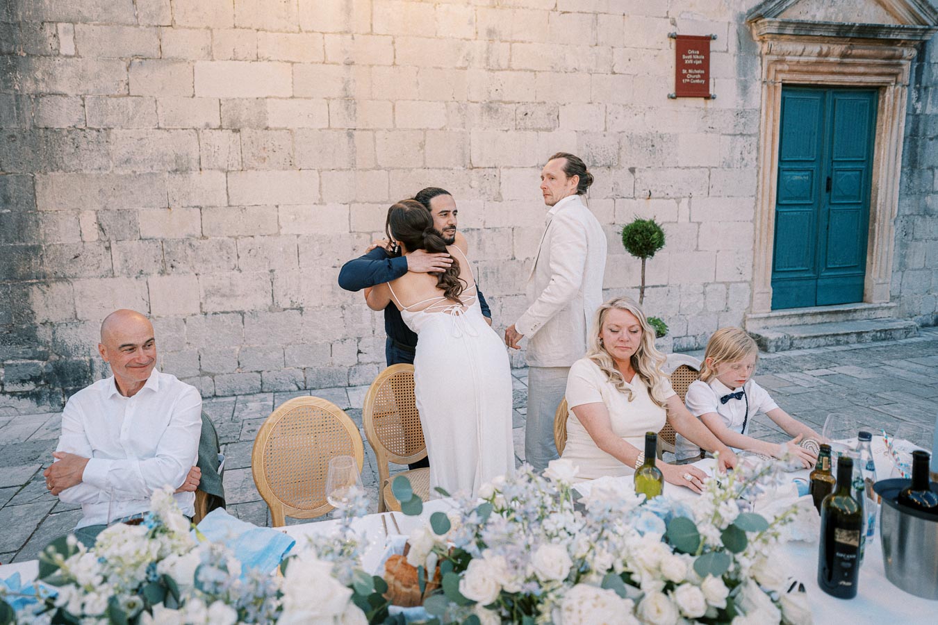 Wedding reception with guests at a beautifully decorated table outdoors, featuring floral arrangements and wine bottles. A woman in a white dress is hugging a man in a suit, while other guests are seated near a historic stone wall.