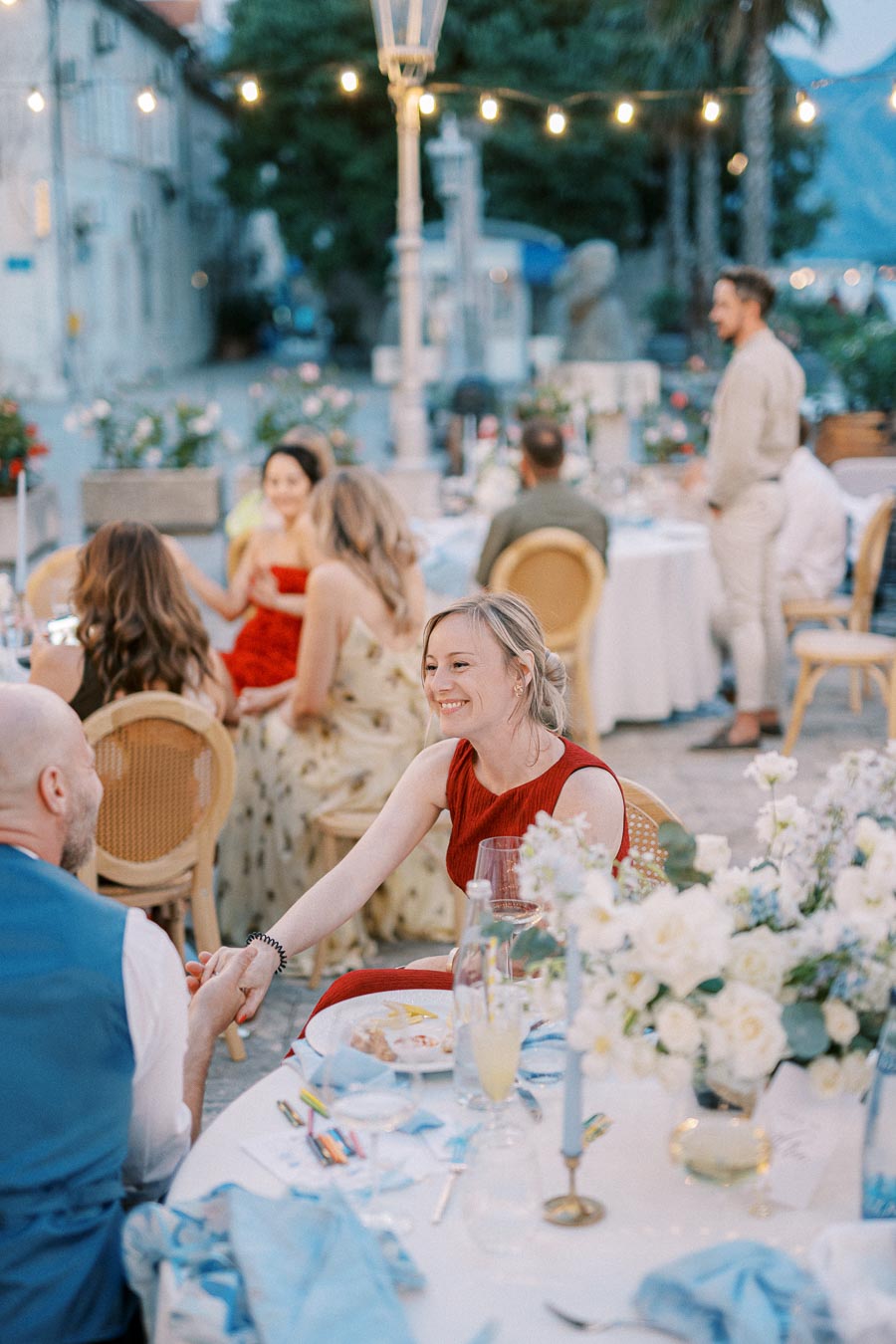 Outdoor dinner event with festive string lights, featuring a woman in a red dress smiling and holding hands with a man across a round table adorned with white flowers and blue accents.