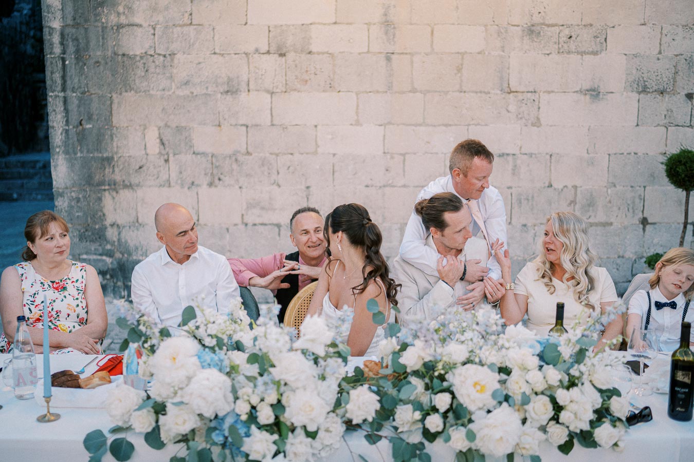 A lively family gathering at a long table decorated with white and blue flowers, featuring people engaged in conversation against a rustic stone wall backdrop, creating a joyful and elegant atmosphere.