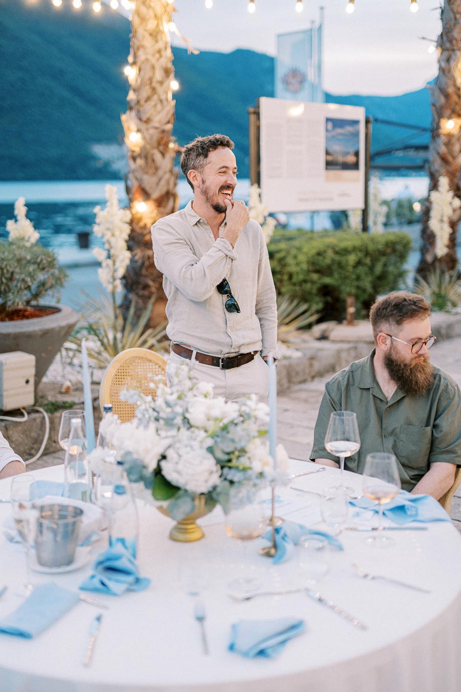 Outdoor dinner gathering with elegantly dressed guests under string lights by the sea, featuring a beautifully set table with blue accents and floral centerpiece.