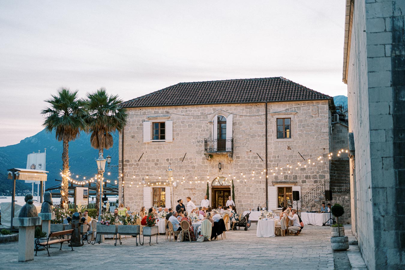 Outdoor evening wedding reception at a rustic stone villa, decorated with string lights and surrounded by palm trees, with guests dining and celebrating in a picturesque courtyard.