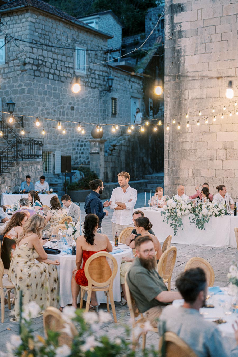 Outdoor evening wedding reception with guests sitting at decorated tables and string lights overhead, set against a stone building backdrop.