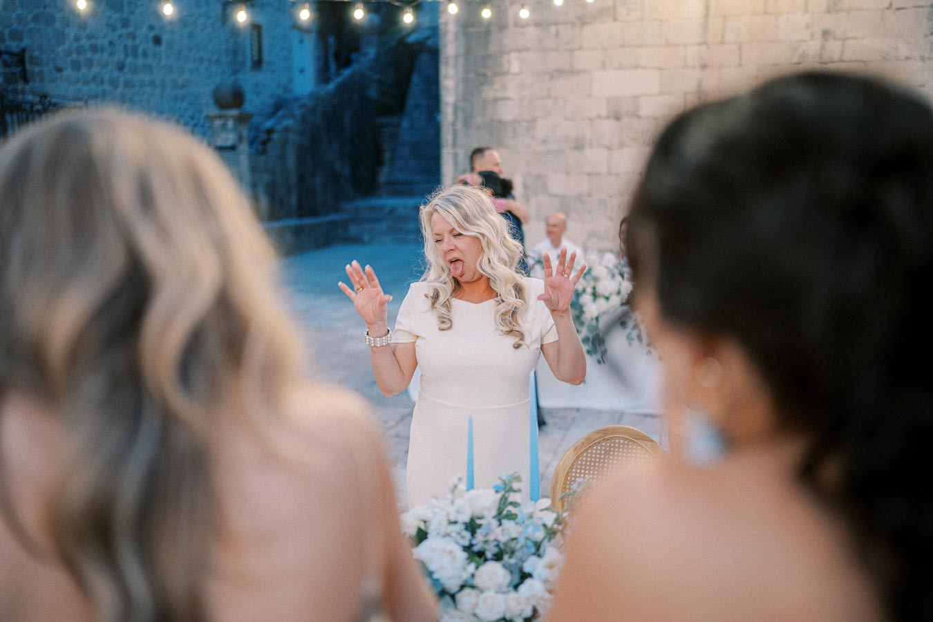 A joyful woman making a playful expression at an outdoor event, surrounded by beautifully arranged flowers and warm evening lights against a rustic stone backdrop.