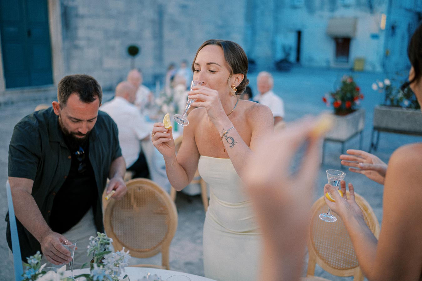 A group of people enjoying a celebration outdoors, with a focus on a woman in a white dress taking a drink from a shot glass while holding a lemon wedge, surrounded by elegantly set tables and other guests.