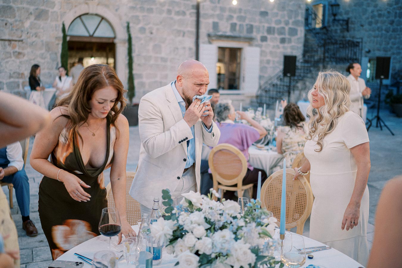 A group of elegantly dressed individuals gather around an outdoor dining table decorated with a floral centerpiece and candles, set in a rustic stone courtyard for a wedding celebration.