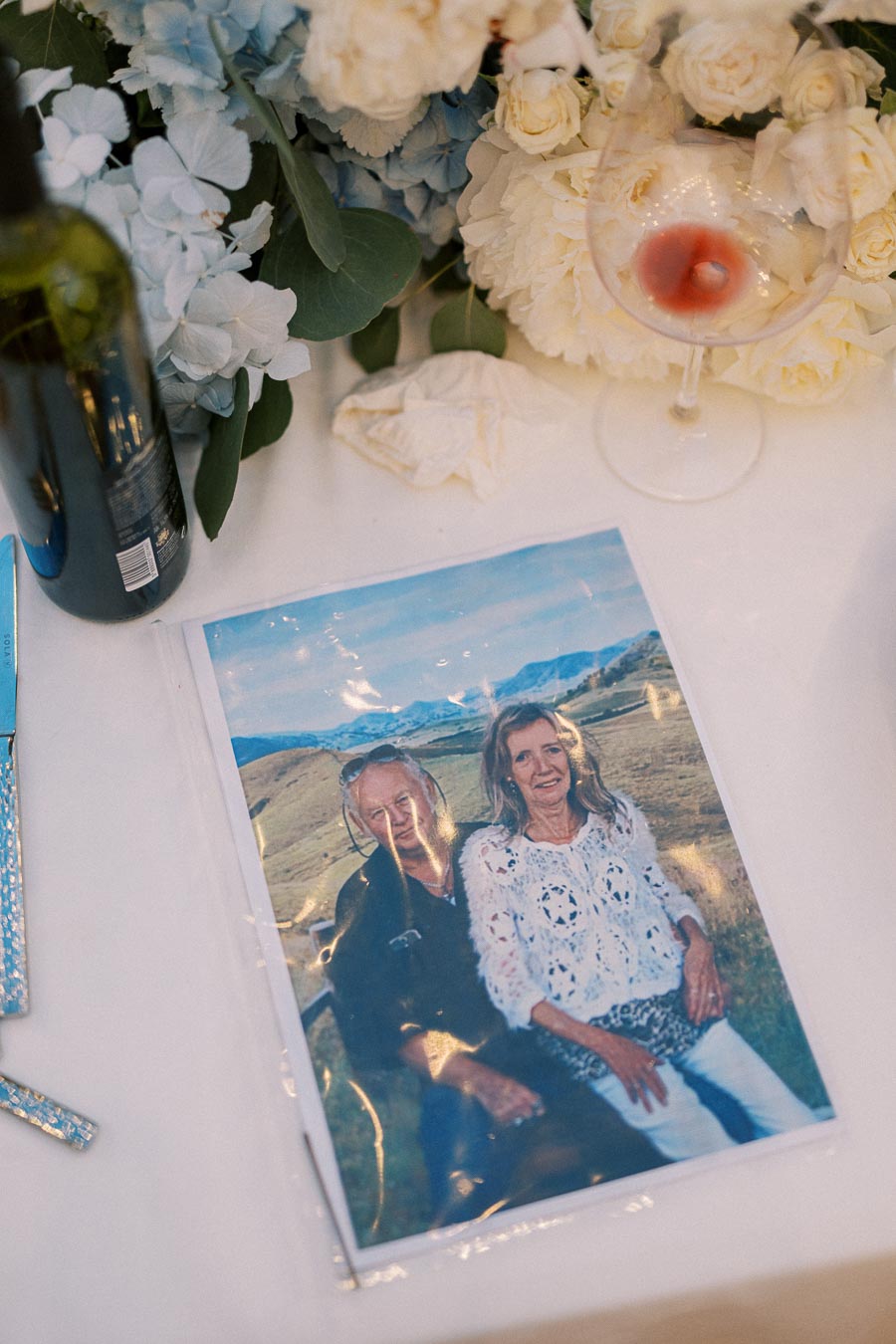 A table setting featuring a photograph of a smiling couple outdoors, surrounded by a wine glass with red wine, a wine bottle, and an arrangement of blue and white flowers.