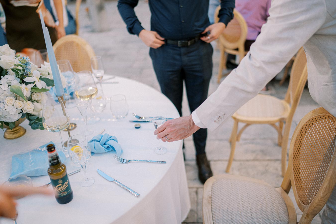Elegant outdoor dining setting with a round table adorned with a white tablecloth, blue napkins, and a floral centerpiece. Guests in formal attire gather around, while one person pours a drink into a wine glass.