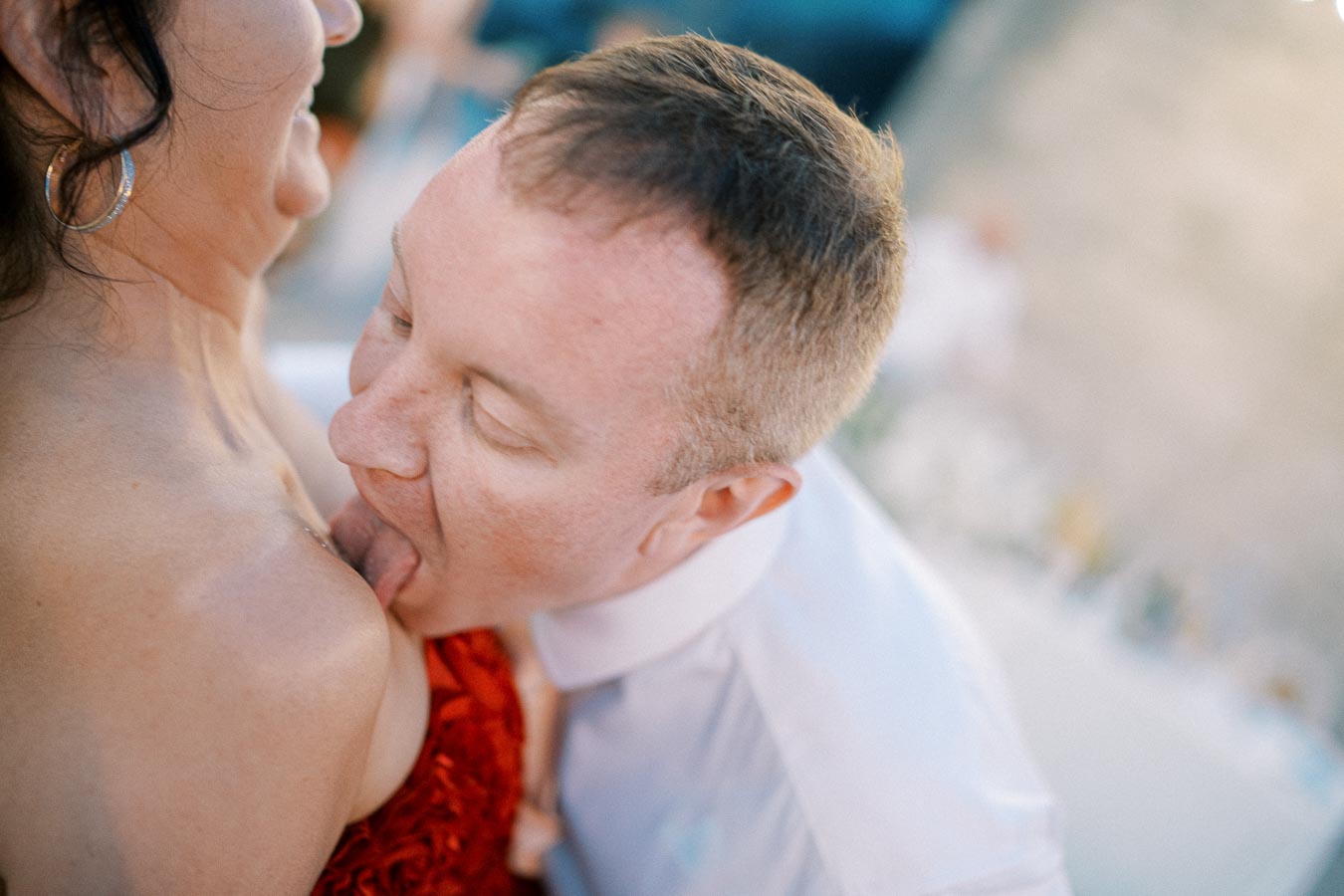 A playful moment captured between two people at a social gathering, with the man playfully licking the woman's shoulder.