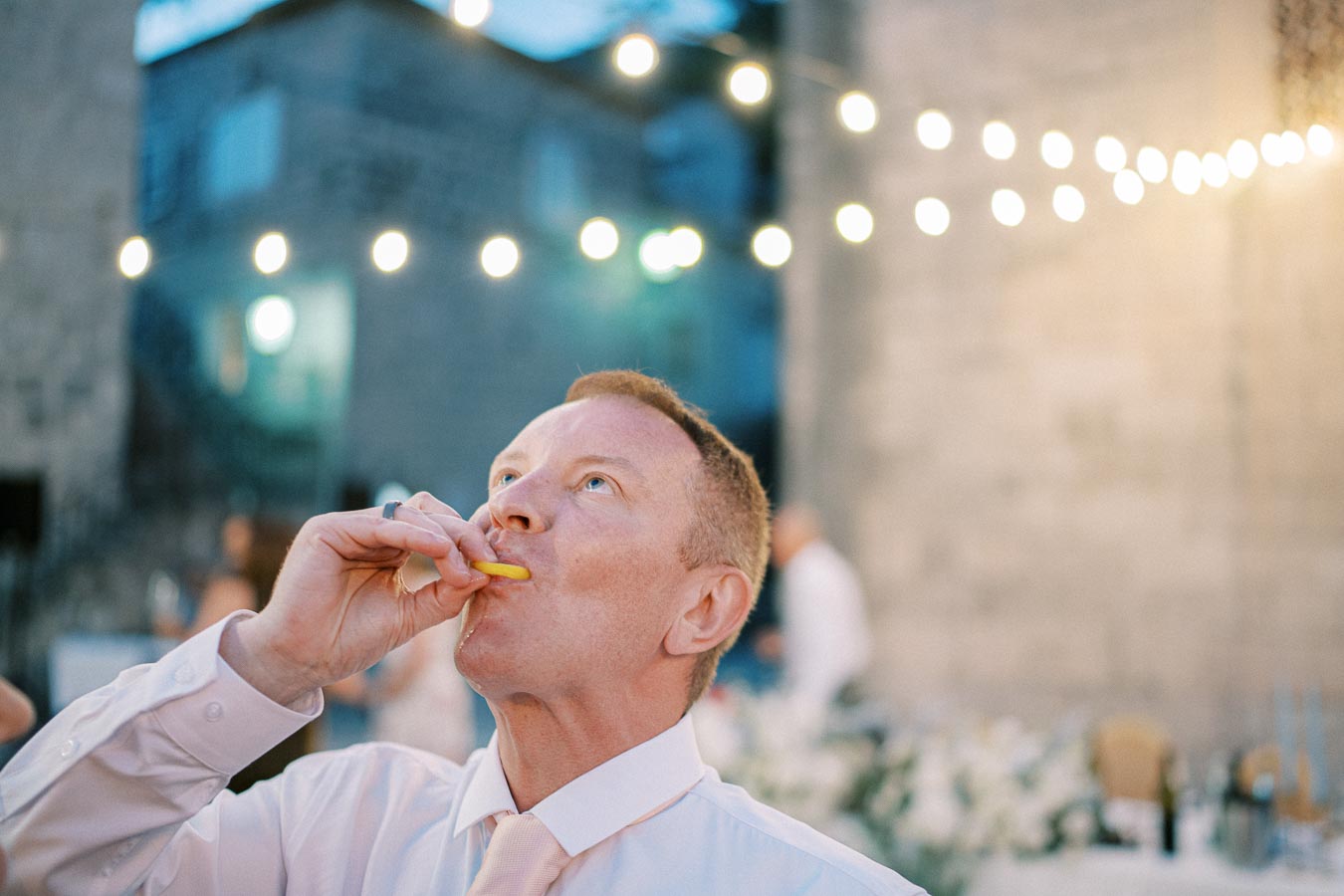 Man enjoying a yellow fruit candy at an outdoor evening event with decorative string lights.