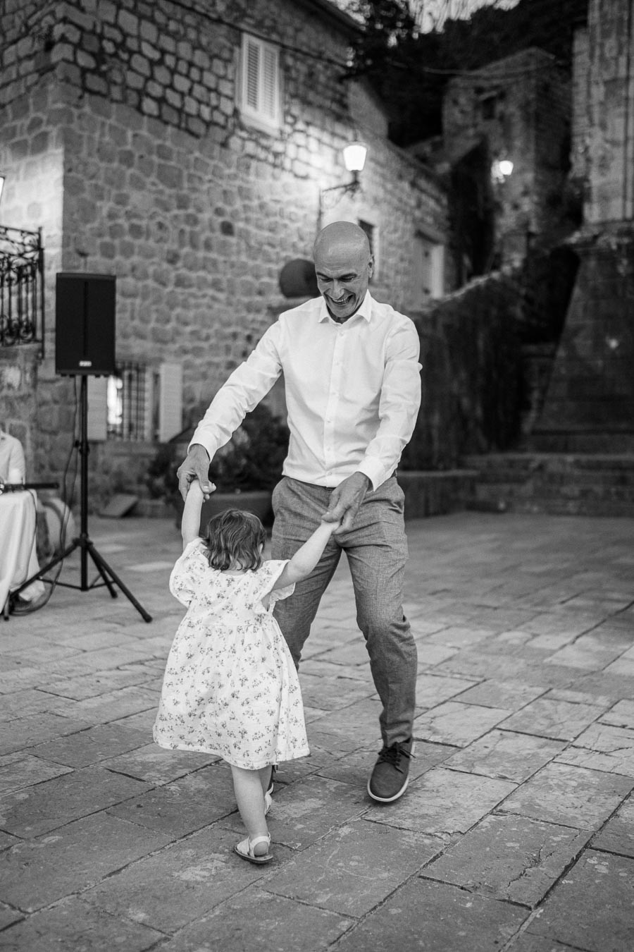 A joyful moment captured in black and white of an older man dancing with a young girl in a floral dress on a cobblestone street, with historic stone buildings in the background.