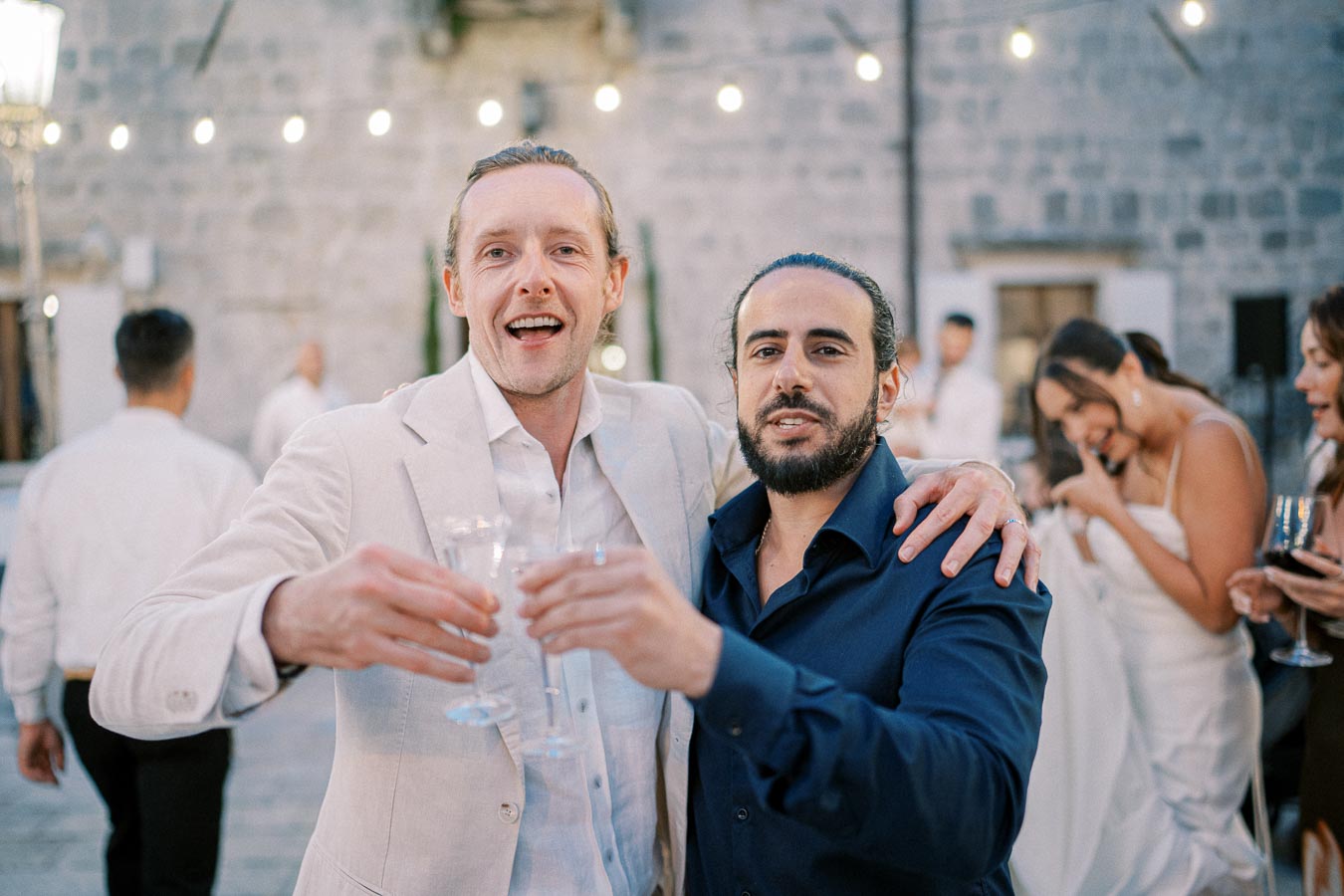 Two friends smiling and raising glasses at a celebration, with people and string lights in the background creating a festive atmosphere.