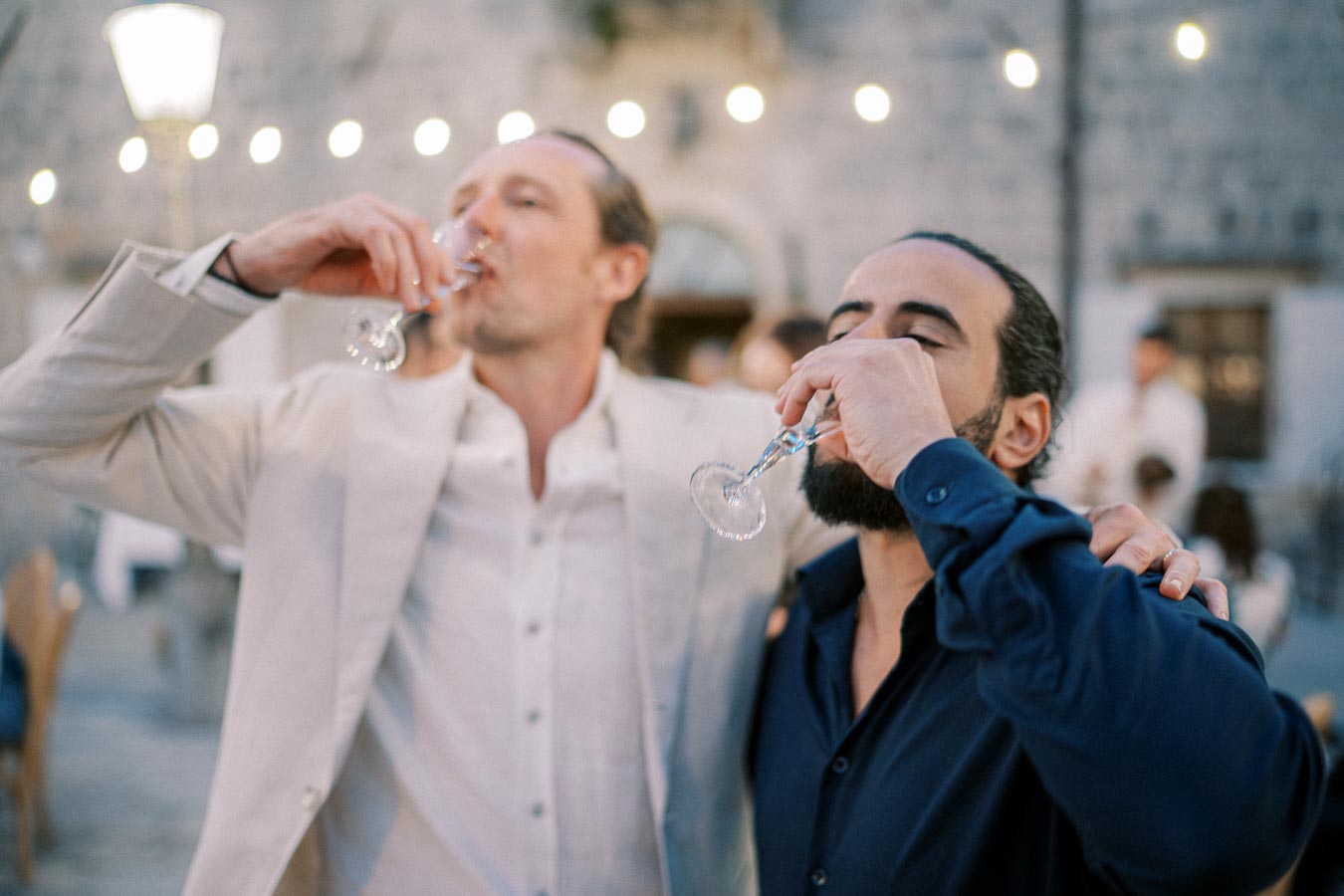 Two people enjoying drinks together at an outdoor social event, with festive lights and a stone wall in the background.