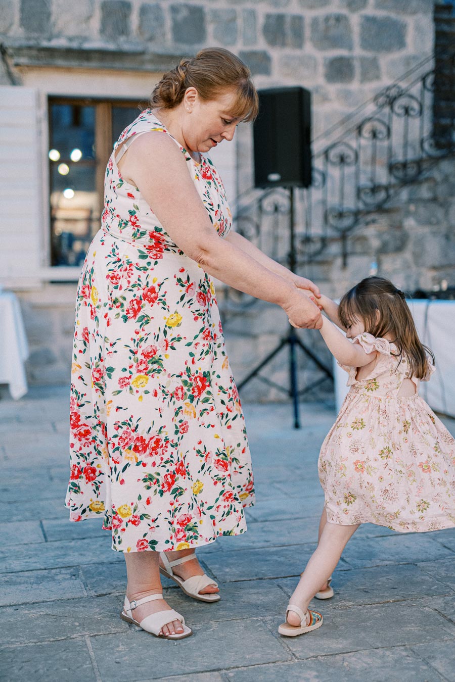 A woman in a floral dress joyfully dances with a young girl in a matching floral outfit on a stone patio, with a rustic stone wall in the background.