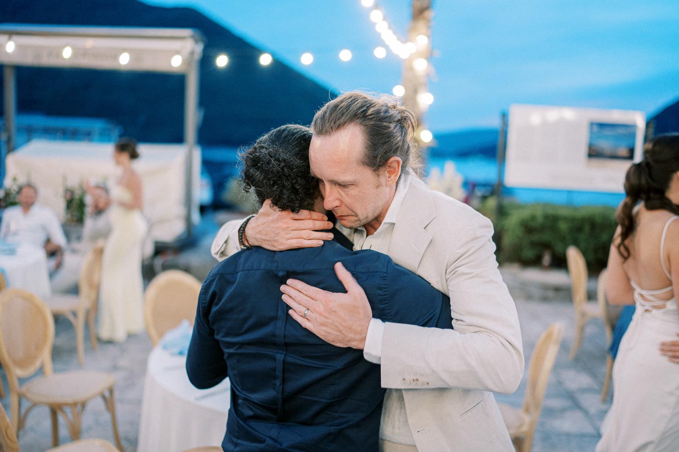Two people hugging at an outdoor event with string lights and mountains in the background, creating a warm and emotional atmosphere.