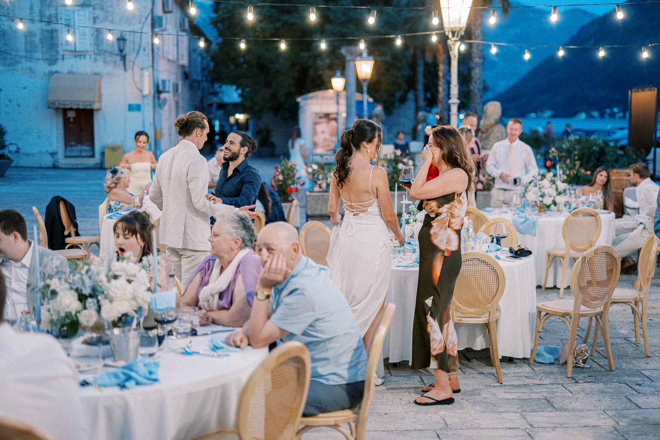 Outdoor evening wedding reception with guests enjoying a lively celebration. Tables are elegantly set with floral arrangements under string lights, creating a festive atmosphere by the waterfront.