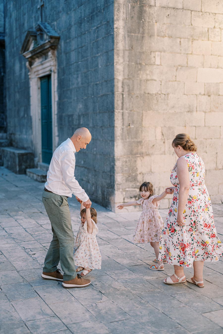 A family enjoying a playful moment on a cobblestone street, with two children in floral dresses being swung by their parents, creating a joyful and lively scene.