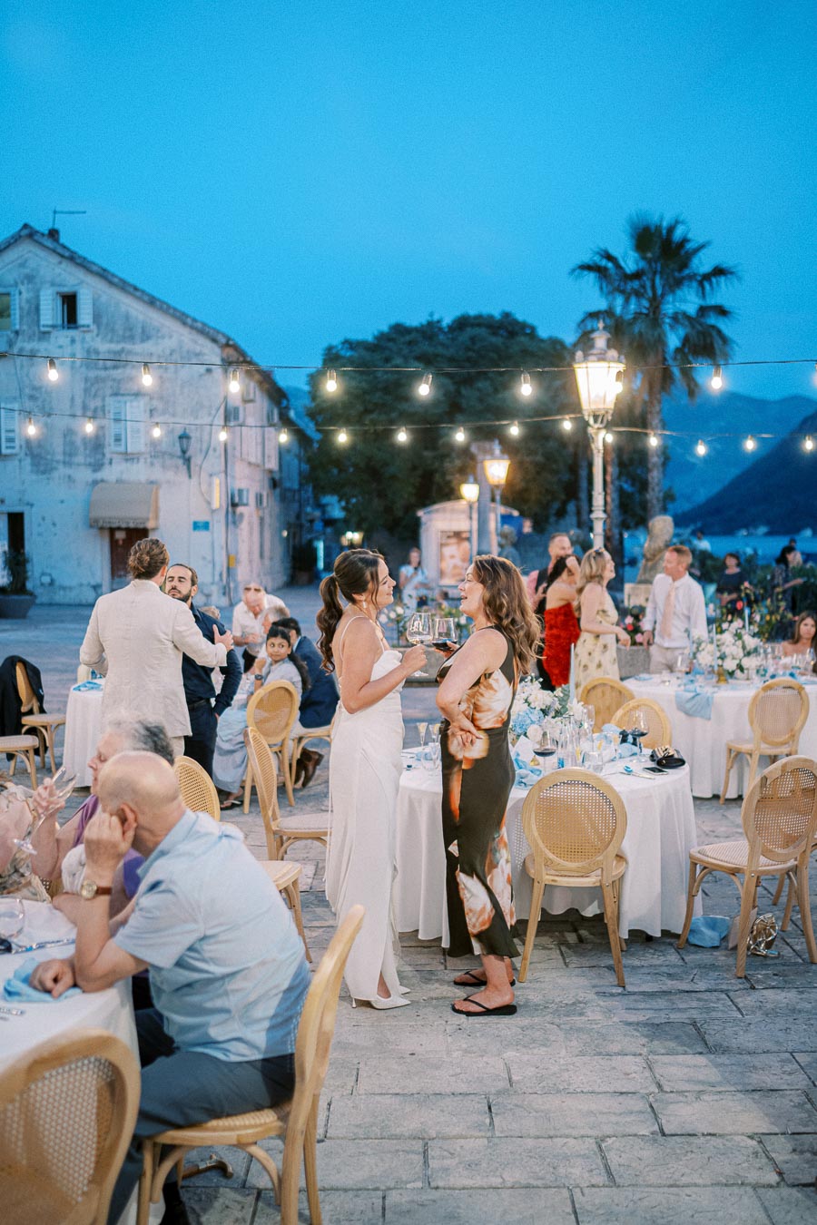 Outdoor evening wedding reception with guests socializing around decorated tables under string lights, set against a backdrop of rustic buildings and palm trees.