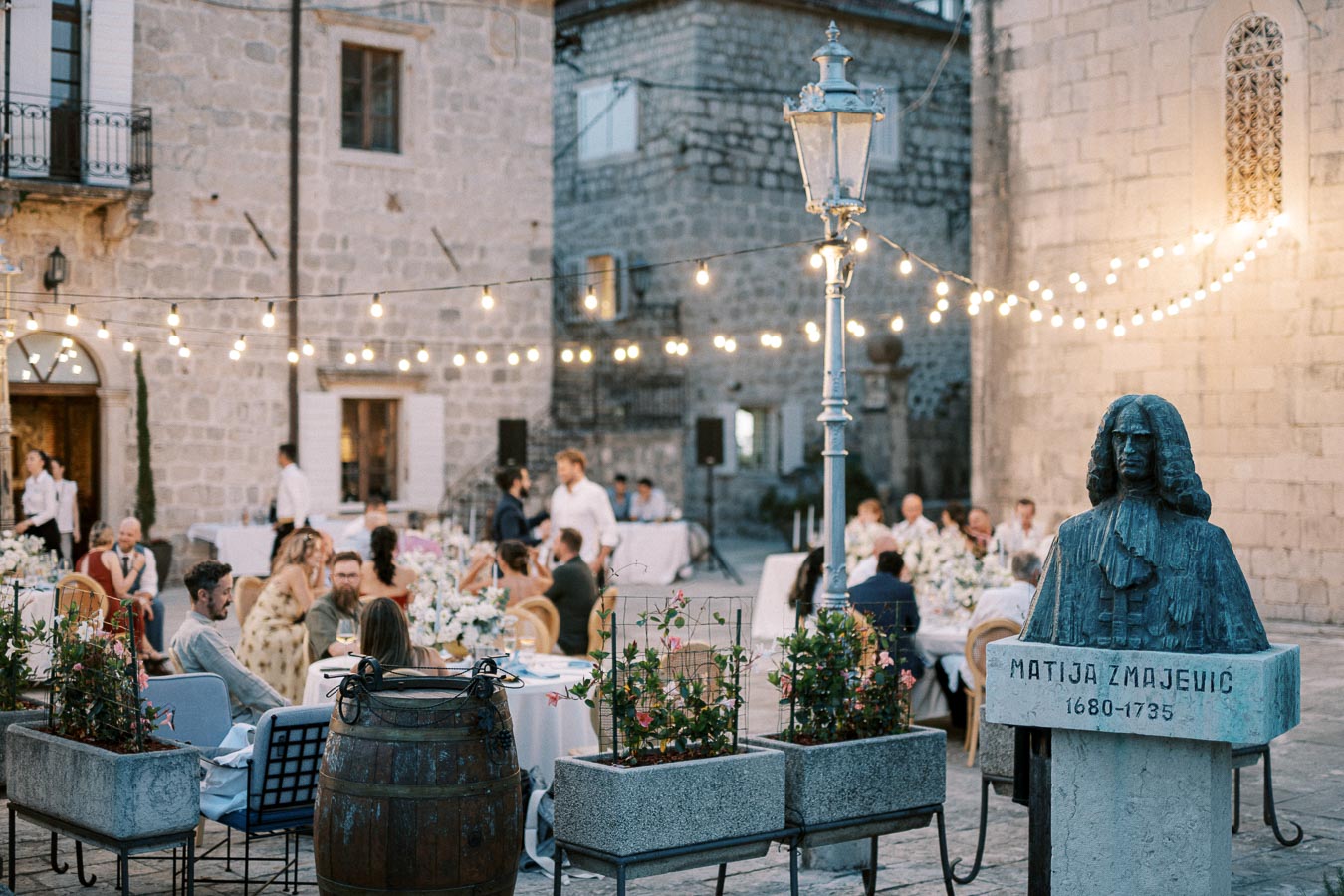 Outdoor event in a historic stone courtyard with string lights, dining guests seated at tables, and a statue of Matija Zmajević in the foreground.
