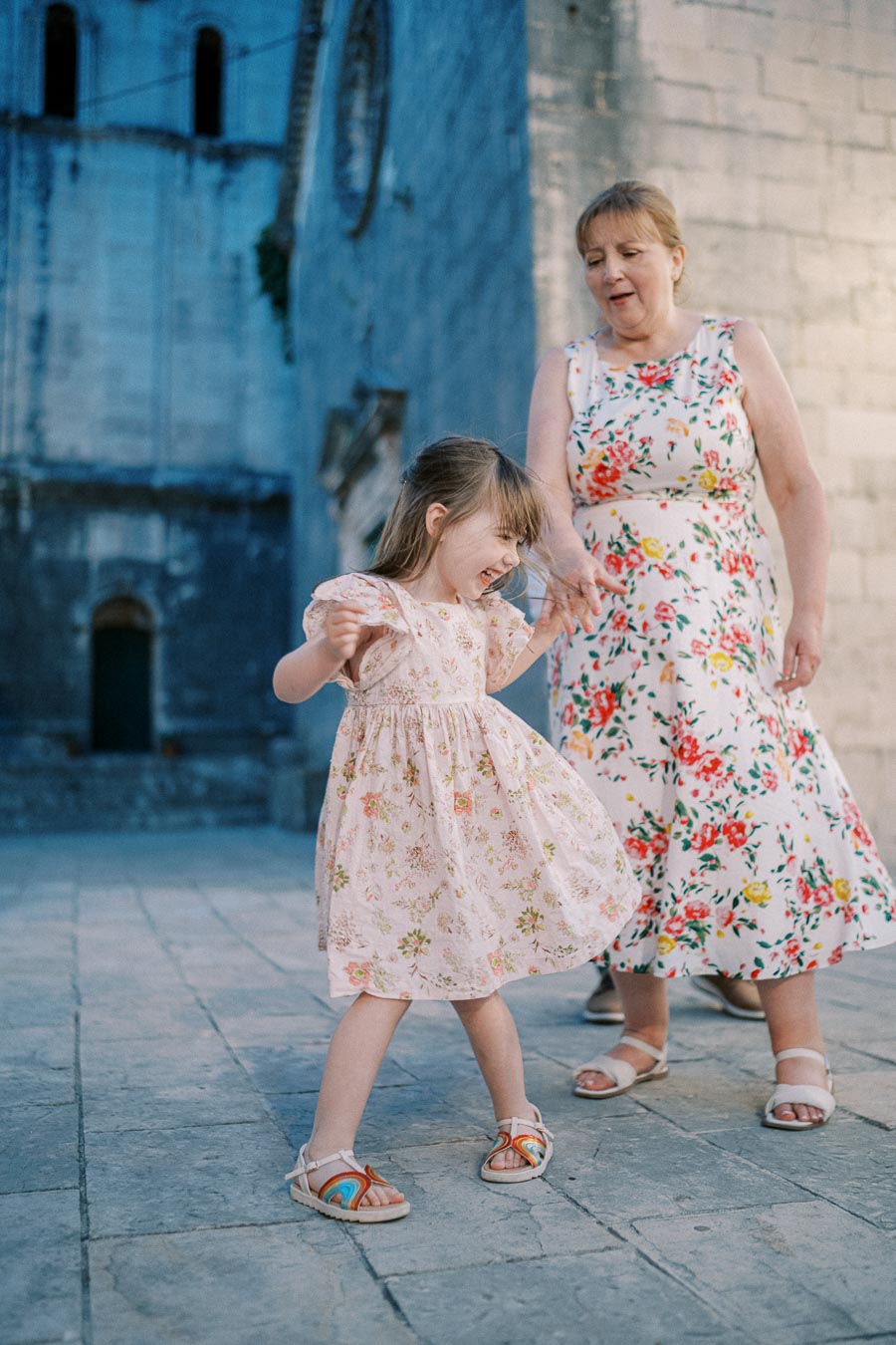 A joyful little girl twirling in a floral dress, holding hands with a woman in a colorful dress, in front of a historic stone building.