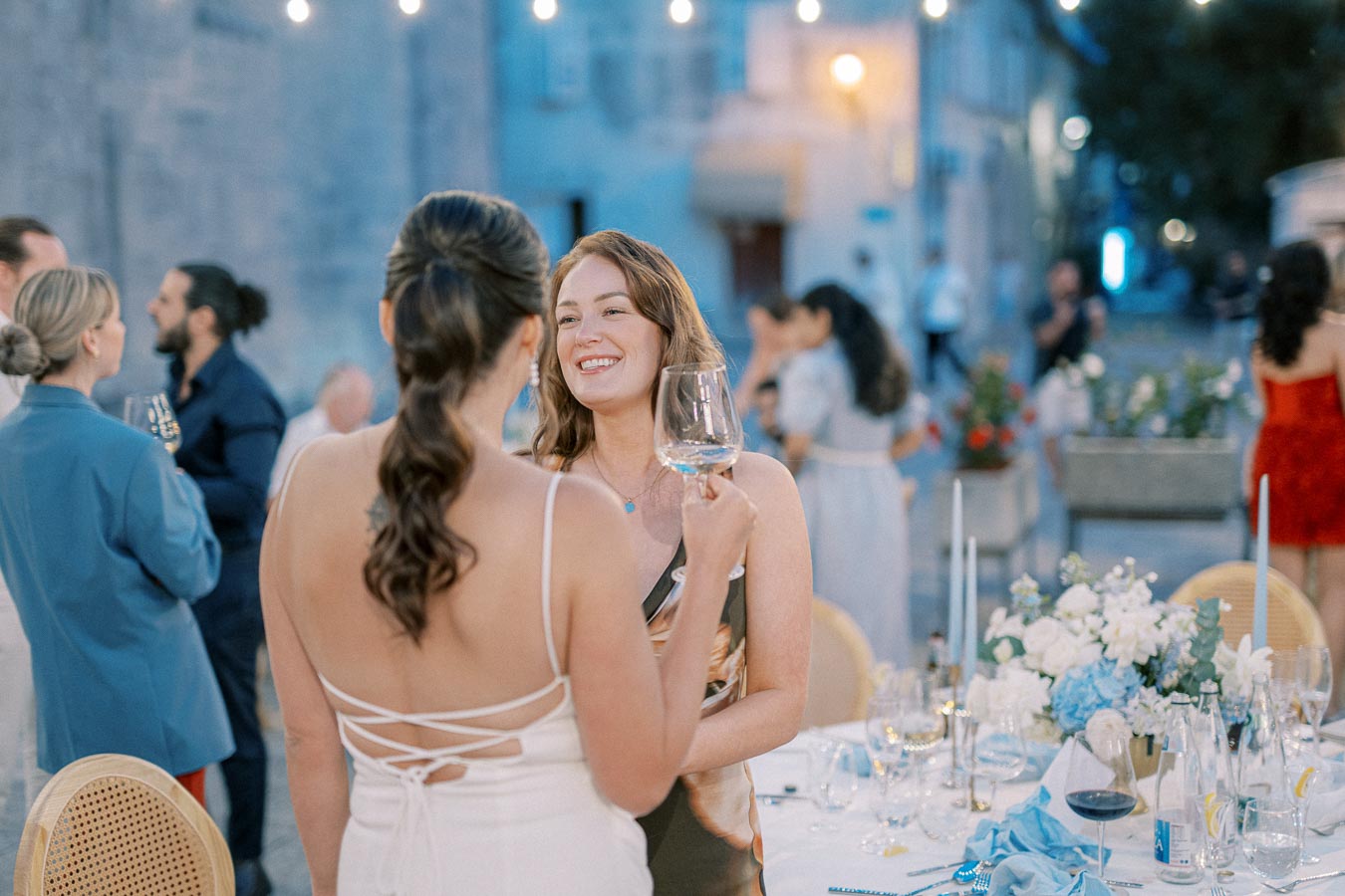 Two women enjoying a conversation at an elegant outdoor event, one holding a wine glass, with ambient lighting and a beautifully decorated table with flowers in the foreground.