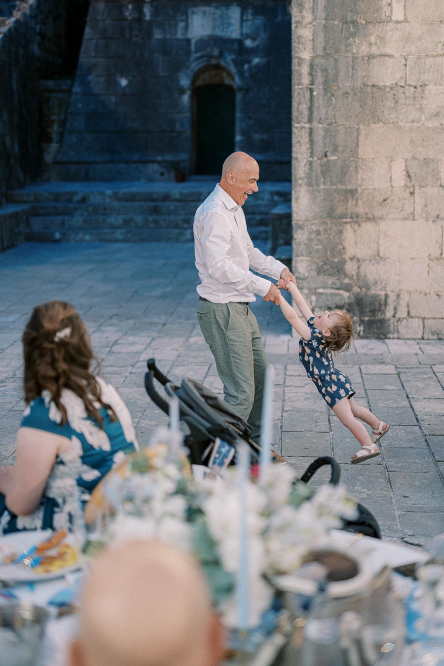 Grandfather joyfully spins granddaughter outdoors at a celebration, with festive decorations and an ancient stone building in the background.