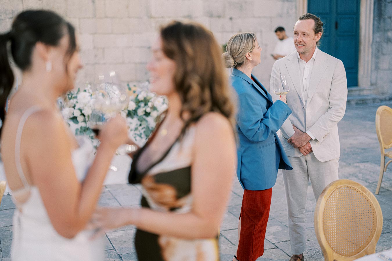 A group of people socializing at an outdoor event, with some holding glasses of wine. Two women in the foreground are blurred, while a man in a white suit and a woman in a blue blazer engage in conversation in the background. The setting features stone walls and elegant floral arrangements.