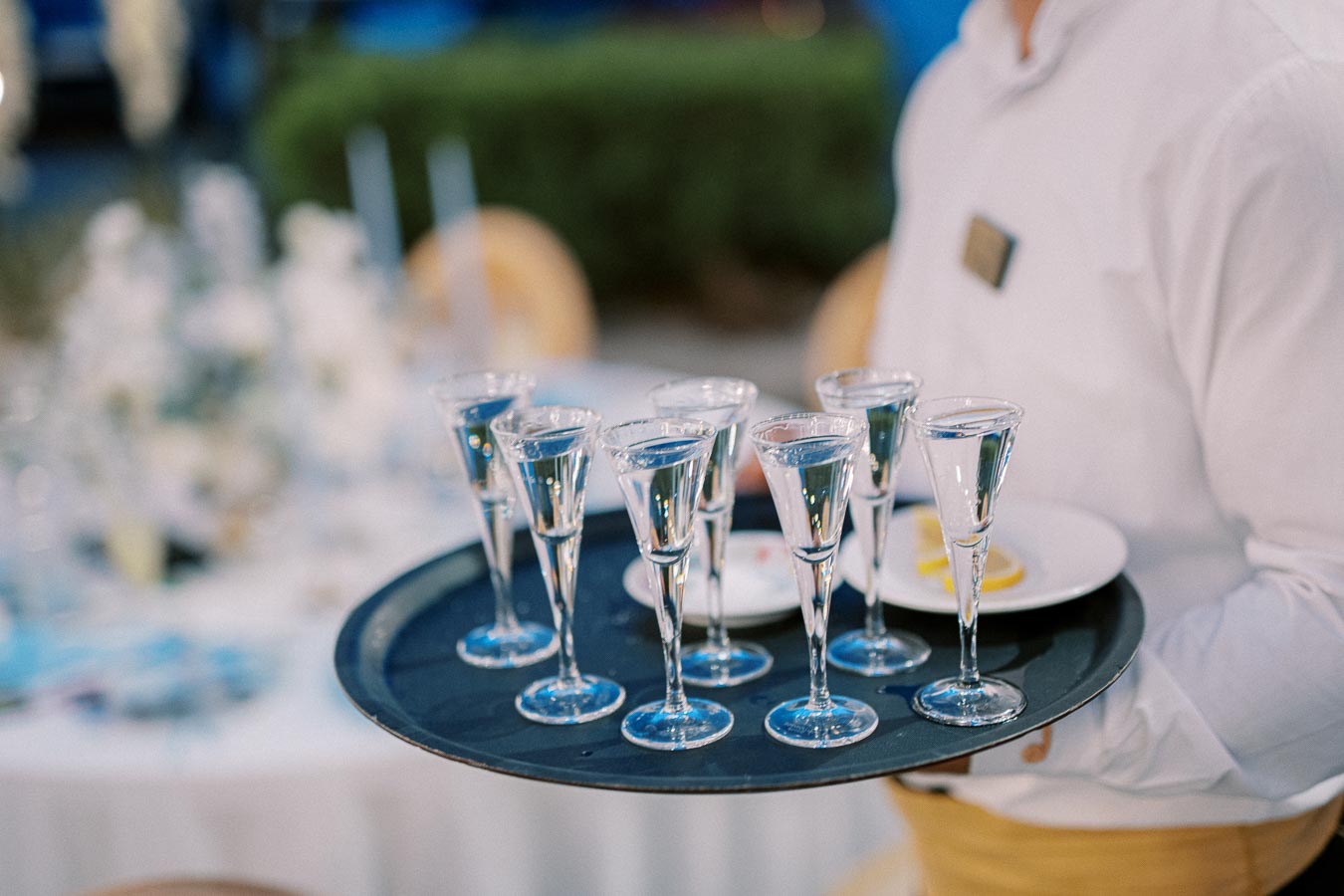 A waiter holding a tray of filled cordial glasses at an elegant outdoor event.